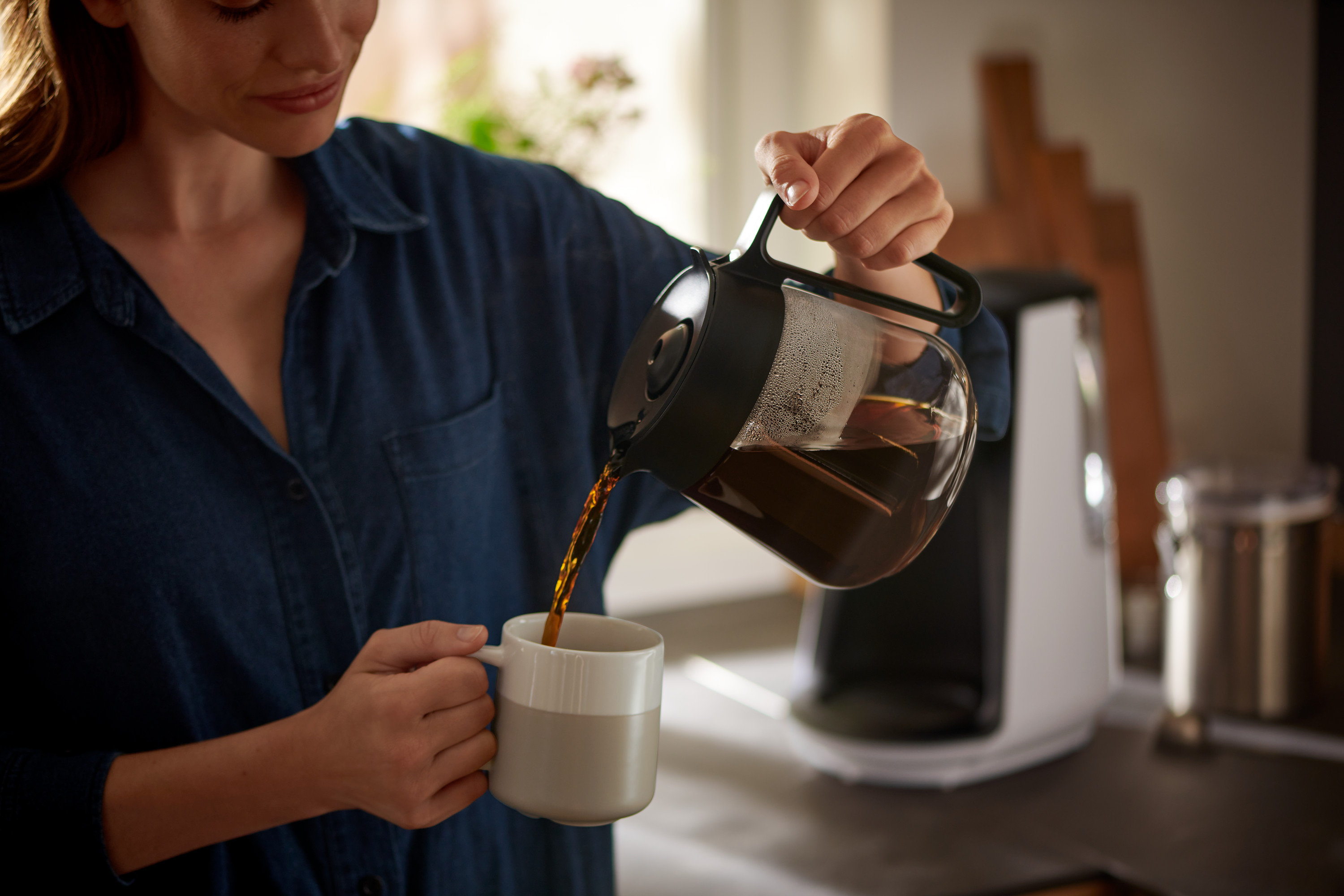 Kadın, bir demlikten bir fincana kahve dolduruyor. Arka planda »Let's Brew« Filtre Kahve Makinesi görülüyor.