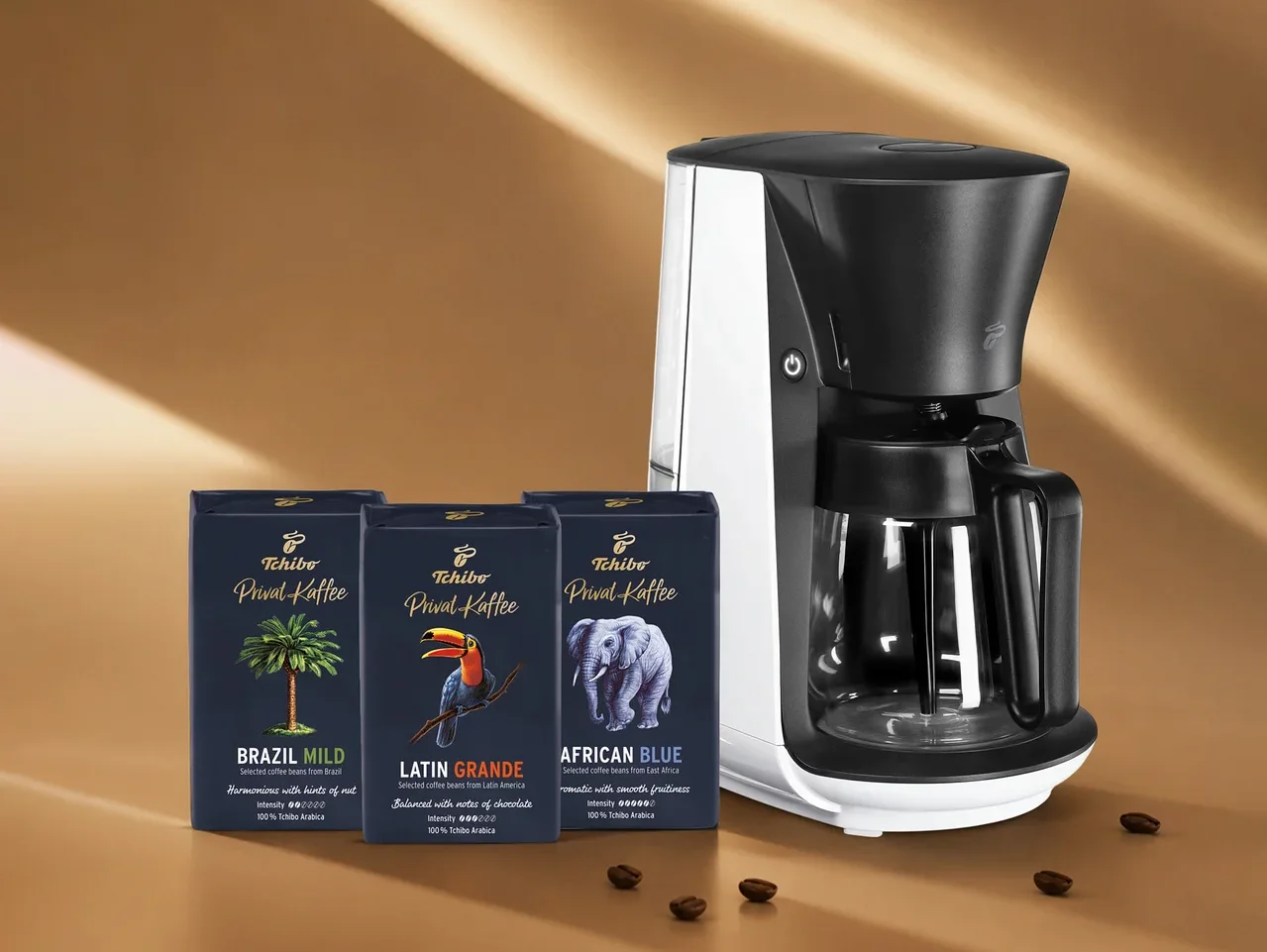 Beyaz ve siyah bir kahve makinesi, palmiye ağacı, tukan ve fil motifli üç koyu mavi Tchibo Privat Kaffee paketiyle birlikte kahverengi bir zeminde, etrafa saçılmış kahve çekirdekleriyle duruyor.