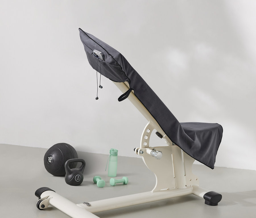 Bir ağırlık sehpası gri bir bezle örtülüdür. Sehpanın yanında bir sağlık topu, bir kettlebell, bir su şişesi ve dambıllar bulunmaktadır.