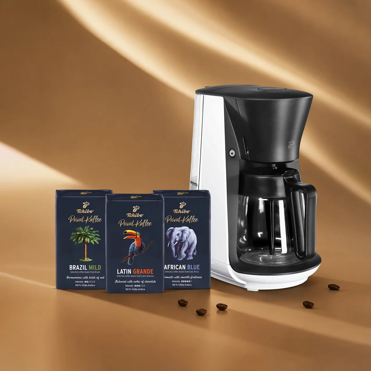 Beyaz ve siyah bir kahve makinesi, palmiye ağacı, tukan ve fil motifli üç koyu mavi Tchibo Privat Kaffee paketiyle birlikte kahverengi bir zeminde, etrafa saçılmış kahve çekirdekleriyle duruyor.